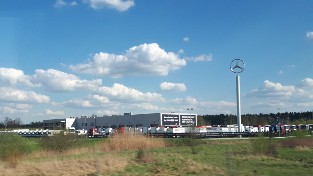 Mercedes-Benz-Fabrik in Deutschland mit Fahrzeugen auf der Straße, Grünflächen, Gebäuden und einem Logo-Pfahl unter einem bewölkten Himmel.