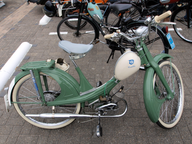 Ein grünes Moped mit weißem Rahmen und schwarzem Sitz neben anderen Motorrädern auf einem Gehweg geparkt, mit ein paar Menschen in der Nähe.