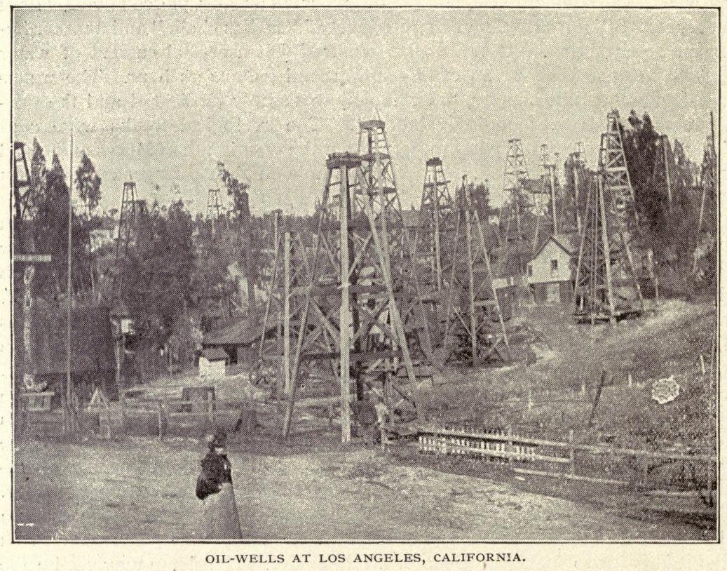Schwarz-weiß-Foto einer Ölquelle in Los Angeles, Kalifornien, mit einer Person zwischen Pfählen, Bäumen, Häusern und einem Zaun, mit Text unten.