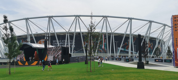Olympiastadion in London mit Menschen, einem Baum im Vordergrund, Bannern am Gebäude und einem bewölkten Himmel.