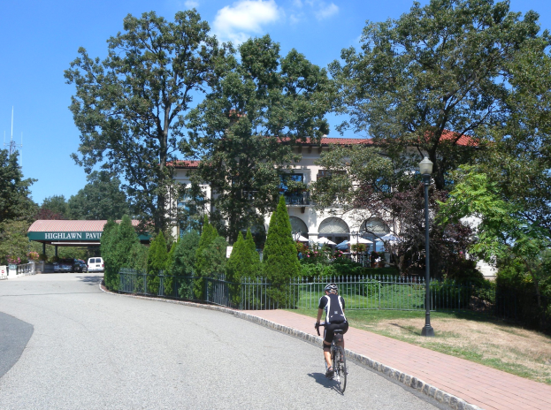 Eine Person mit Helm fährt ein Fahrrad eine von Bäumen gesäumte Straße mit Laternen, Geländern und Gras entlang, mit einem Gebäude, einem Schuppen und einem bewölktem Himmel im Hintergrund.