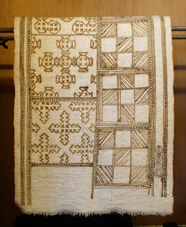 Weißer und brauner Stoffteppich mit detaillierten Designs, der an einer Wand neben einer Holztür hängt.