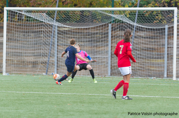 Eine Gruppe von Frauen, die auf einem grünen Rasenfeld umgeben von Bäumen Fußball spielen, mit einem Tor im Hintergrund und Text am unteren Rand des Bildes.