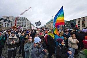 Eine große Gruppe von Menschen steht vor einem Gebäude und hält Fahnen und Transparente mit Texten, darunter eines mit der Aufschrift "Lgbtq+ Rechte Demonstration in Berlin", einige tragen Mützen und Taschen; im Hintergrund sind Gebäude, ein Kran und ein bewölkter Himmel zu sehen.