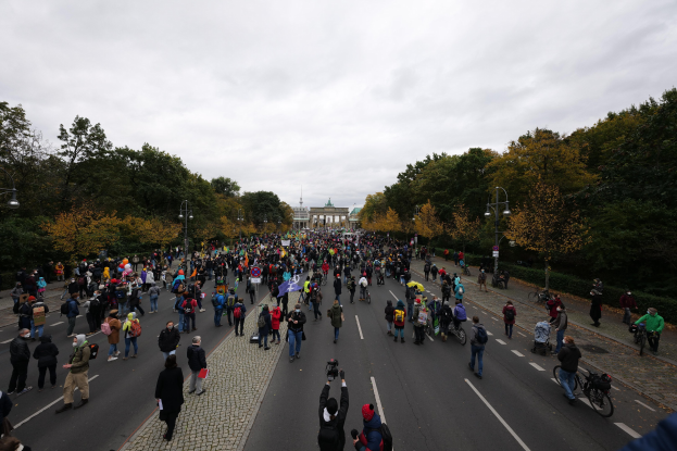 Eine große Gruppe von Menschen marschiert eine baumbestandene Straße mit Laternenpfählen entlang, mit Kameras in der Hand, mit einem Gebäude und einem klaren Himmel im Hintergrund.