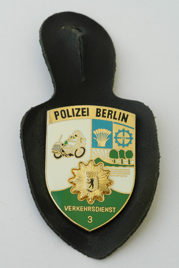 Ein Polizeistern mit der Aufschrift "Polizei Berlin" darauf, mit einem blauen Schild und einem weißen Stern in der Mitte, umgeben von einem weißen Rand, auf einer Oberfläche plaziert.