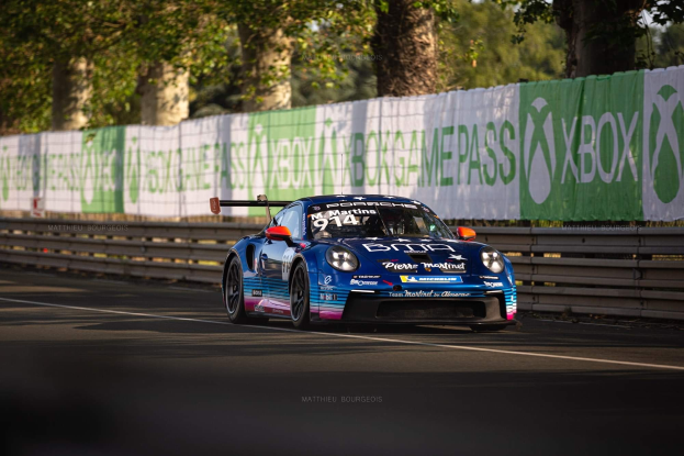 Blauer Porsche 911 GTS auf einer Rennstrecke mit einem Zaun und Bannern im Hintergrund, Bäume und einen klaren Himmel.