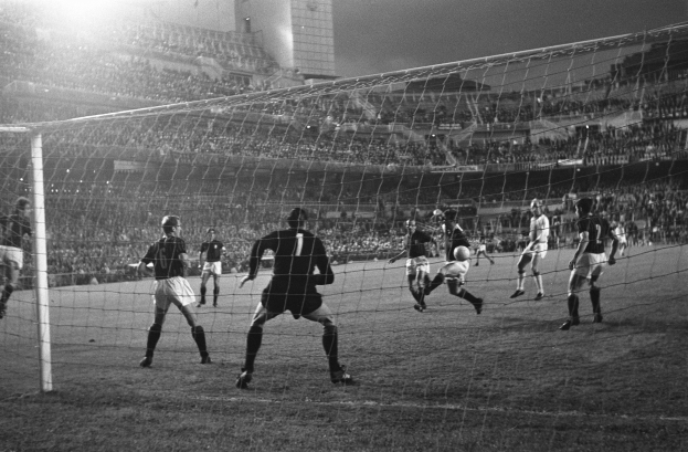 Eine Gruppe von Männern, die Fußball spielen, auf einem Stadionrasen mit einem Torpfosten im Vordergrund und einem Gebäude im Hintergrund, dargestellt in Schwarz-Weiß.