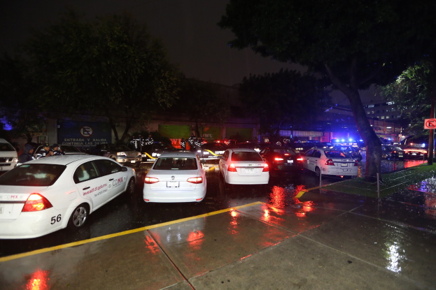 Eine Gruppe von Polizeiwagen auf einem Parkplatz bei Nacht, mit einem Schild, Bäumen, Gebäuden, einer Fahne und einem bewölkten Himmel.