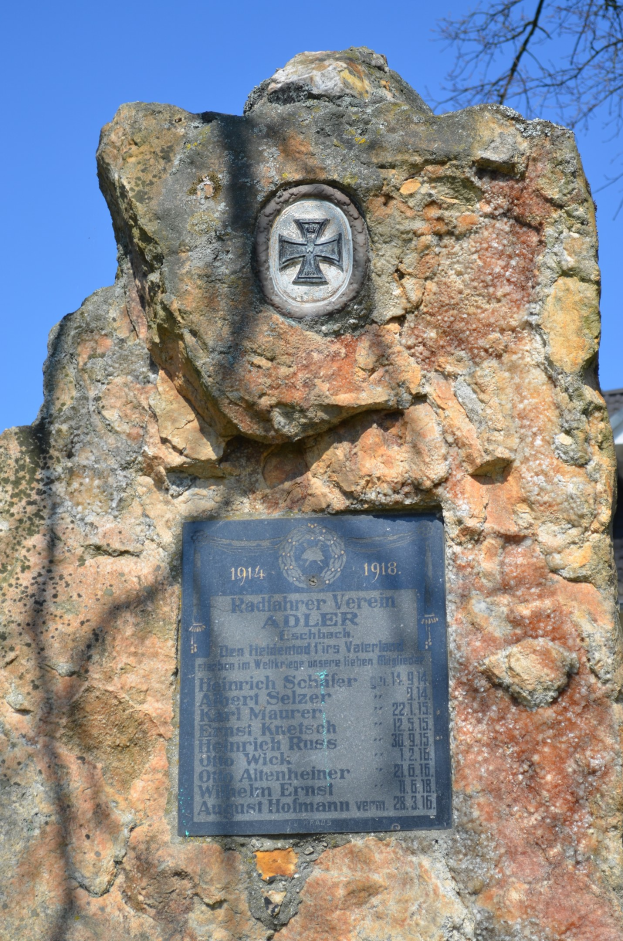 Denkmalstein mit einer Plakette vor einem Gebäude, Baum im Hintergrund und Himmel darüber.
