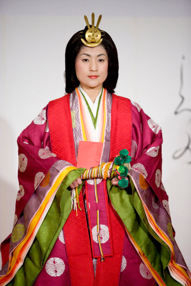 Eine Frau in einem traditionellen japanischen Kimono, das ein leuchtend rotes und grünes Kleid mit aufwändigen Mustern zeigt, steht selbstbewusst auf einem Laufsteg, ihr Haar ist zu einem ordentlichen Knoten gebunden.