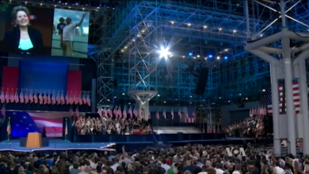 Eine große Menschenmenge sitzt vor einer Bühne mit einem Rednerpult, einem Mikrofon, Flaggen, einem Bildschirm und Beleuchtungsausrüstung und wartet auf eine Debatte.