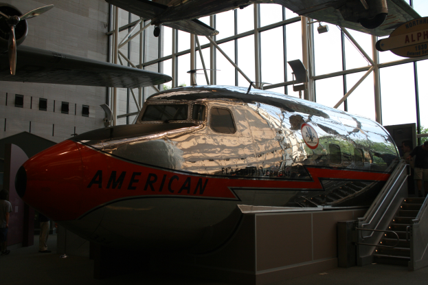 Ein American-Airlines-Flugzeug in einem Museum mit Menschen drumherum, Treppe führt rechts zum Flugzeug hoch und Gebäude, Bäume und klarer blauer Himmel im Hintergrund.