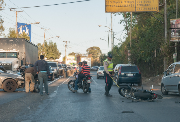 Eine Gruppe von Menschen steht um ein verunglücktes Motorrad auf der Straße herum, umgeben von mehreren Fahrzeugen, darunter ein Lastwagen, sowie Hintergrundelementen wie Bäumen, Pfählen, Laternen und Schildern.