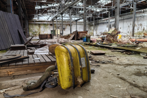 Ein gelber Koffer steht auf einem Holzboden in einem verlassenen Gebäude, umgeben von verstreuten Gegenständen wie Holzplanken und Papieren, mit einer Wand, Pfählen und einem Dach mit Metallstangen im Hintergrund.