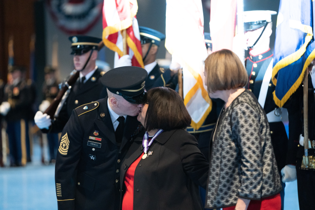 Zwei Menschen umarmen und küssen sich, während eine Gruppe in Uniformen und Mützen, einige mit Fahnen, in der Nähe steht.