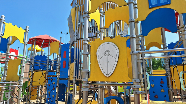 Ein Spielplatz mit bunten Spielgeräten, darunter Stangen, Pfosten und ein Schirm, einige Menschen, Bäume im Hintergrund und ein klarer blauer Himmel.