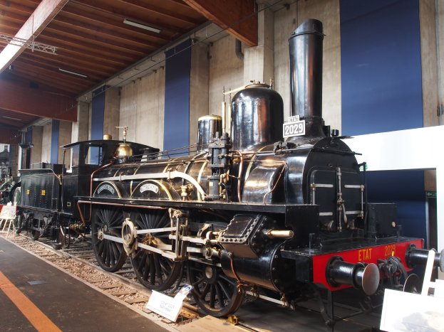 Eine gro├če, alte Dampflokomotive in einem Museums Setting mit umliegenden Schienenwegen, Informationsschildern und anderen Ausstellungen, vor einer Wand mit Deckenbeleuchtung.