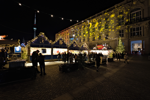 Ein lebendiger Weihnachtsmarkt in Berlin, Deutschland, mit Menschen, die sich um hell erleuchtete und geschmückte Stände drängen, vor Gebäuden mit Fenstern unter einem dunklen Himmel.