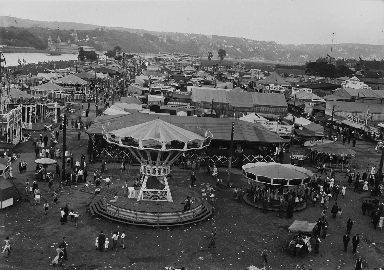 Ein altes Schwarz-Weiß-Foto eines pulsierenden Volksfestes mit einem Karussell in der Mitte, umgeben von Menschen, Buden, Polen, Bäumen, Gebäuden und einer Brücke im Hintergrund unter einem Himmel.
