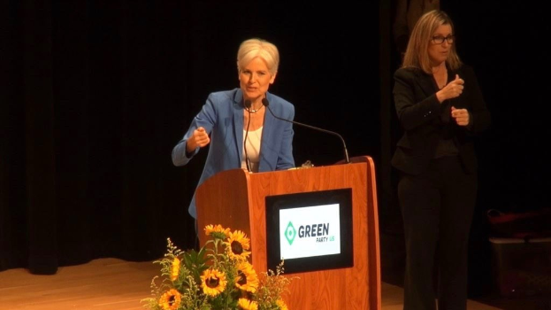 Zwei Frauen stehen an einem Podium mit einem Mikrofon, Bildschirm und Blumen und sprechen auf einer Grünen Party-Veranstaltung zum Thema Umwelt nachhaltigkeit.