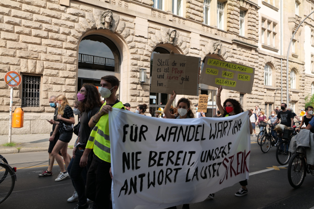 Eine Gruppe von Demonstranten marschiert durch eine Straße in Berlin, einige halten Schilder und andere fahren Fahrräder, mit einem historischen Gebäude mit gewölbigen Fenstern, Säulen und Skulpturen im Hintergrund.