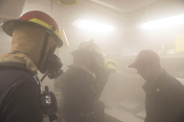 Feuerwehrleute mit Helmen und Rauch, der aus ihren Mündern kommt, einer hält eine Kamera, während einer Übung mit Hintergrundbeleuchtung und -objekten.