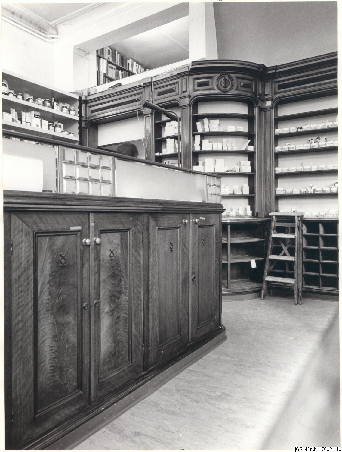 Schwarz-weiß-Foto eines vintage-Apothekeninneren mit einer Theke, Schränken, einer Leiter, Regalen voller Behälter und Flaschen und Text oben.