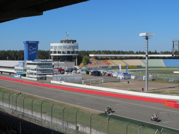 Gruppe von Motorradfahrern auf einer Rennstrecke, umgeben von einem Zaun, Fahrzeugen, einem Gebäude, Schildern, Pfosten, einem Stadion, Laternen, Bäumen und einer bewölkten Himmel.