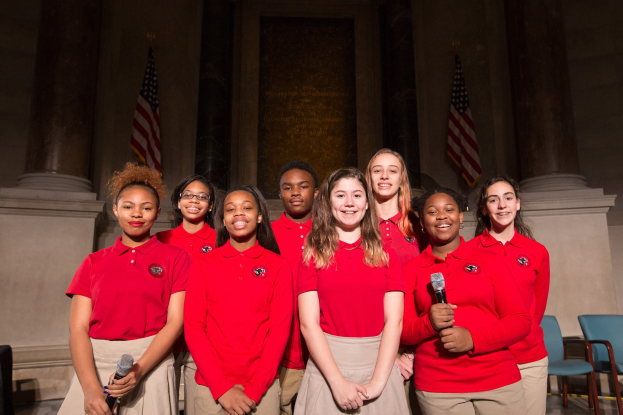 Gruppe junger Frauen in roten Shirts, die strahlend Mikrofone halten und vor Stühlen, Säulen, Flaggen und einer Wand stehen.