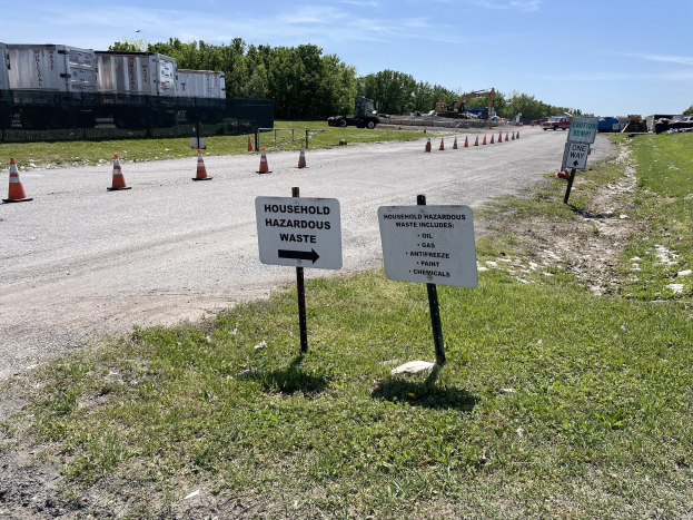 Straßensammelstelle für "Haushaltsgefährliche Abfälle" mit Verkehrskegeln markiert, umgeben von Gras, Fahrzeugen, Bäumen und einem bewölkten Himmel.