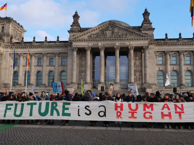 Gruppe von Menschen mit einem Transparent 'Zukunft ist ein Menschenrecht' vor dem Reichstagsgebäude in Berlin, Deutschland, mit sichtbaren Fenstern, Säulen und Bögen des Gebäudes, umgeben von Fahnenmasten und einer bewölkten Himmel.