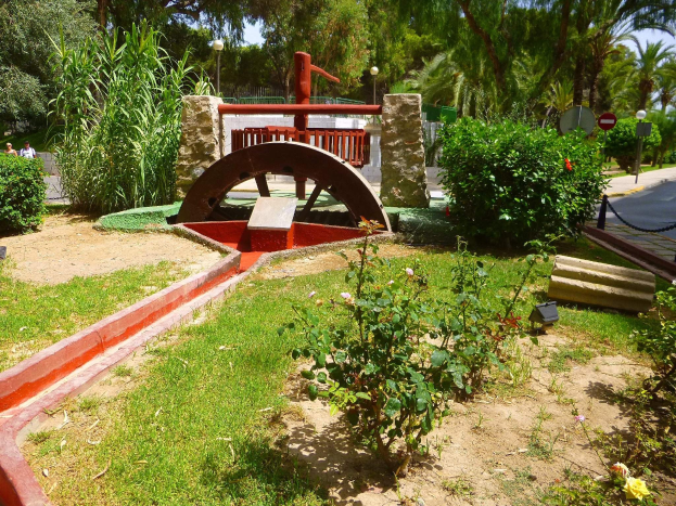 Minigolfanlage in einem Park mit Grünfläche, einer Straße mit Fußgängern, Hinweispfählen und einem klaren blauen Himmel im Hintergrund.