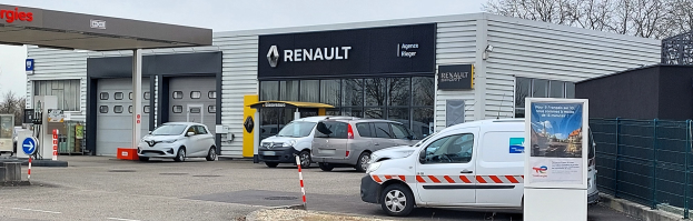 Ein Renault-Autohaus mit parkenden Autos vorne, umgeben von einem Metallzaun, einer Tafel, Pfosten, einem Schild, Bäumen und einem bewölkten Himmel.