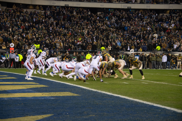 Ein Fußballspiel im Stadion mit Spielern in Sportkleidung und Helmen, umgeben von Zuschauern, Geländern und Schildern.