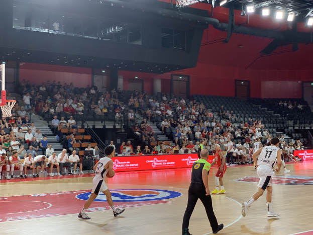 Eine Gruppe von Menschen, die Basketball in einer Turnhalle spielt, mit einem Basketballkorb auf der linken Seite und Zuschauern im Hintergrund.