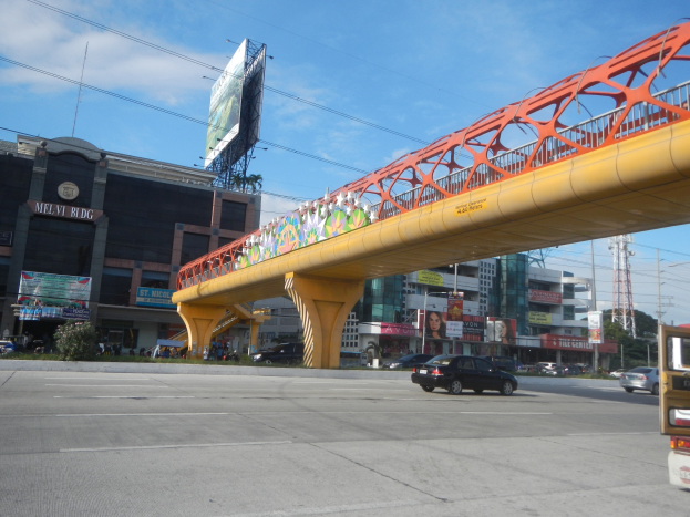 Eine Brücke überspannt eine vielbefahrene Straße mit Autos, umgeben von Gebäuden, Baustellenzäunen, Strommästen mit Käblein, Bäumen und einem bewölkten Himmel.