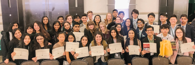 Eine Gruppe von Menschen hält Urkunden in der Hand und posiert für ein Foto bei einer Abschlussfeier, mit Stühlen und einer Wand im Hintergrund.