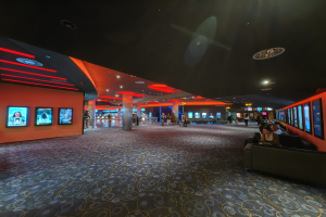 Ein geräumiger Raum mit Menschen auf Sofas sitzend, die Fernsehgeräte an den Wänden anschauen, mit Säulen darin verstreut und Deckenlampen, die den Bereich beleuchten.