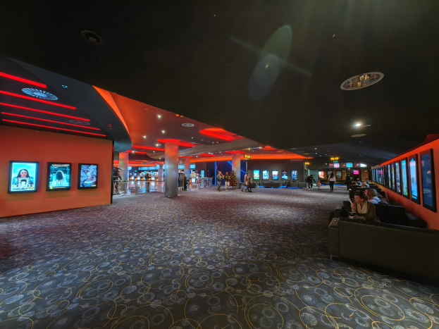 Ein geräumiger Raum mit Menschen auf Sofas sitzend, die Fernsehgeräte an den Wänden anschauen, mit Säulen darin verstreut und Deckenlampen, die den Bereich beleuchten.