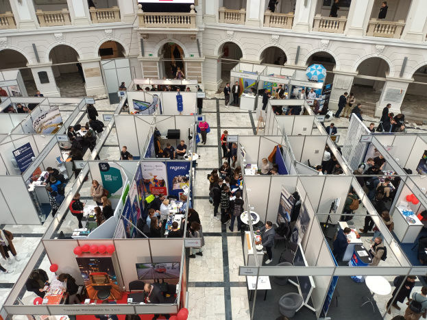 Eine große Gruppe von Menschen auf einer internationalen Fachmesse mit Ständen, Bannern, Tischen, Stühlen und Luftballons vor einem Gebäude mit Bögen, Säulen, Geländern und einem Bildschirm.