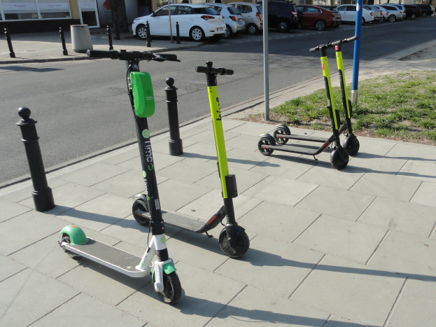 Drei Elektro-Scooter auf der Straße geparkt, umgeben von Gras, Pfählen, Fahrzeugen, Gebäuden mit Fenstern, Bäumen und einem klaren blauen Himmel.