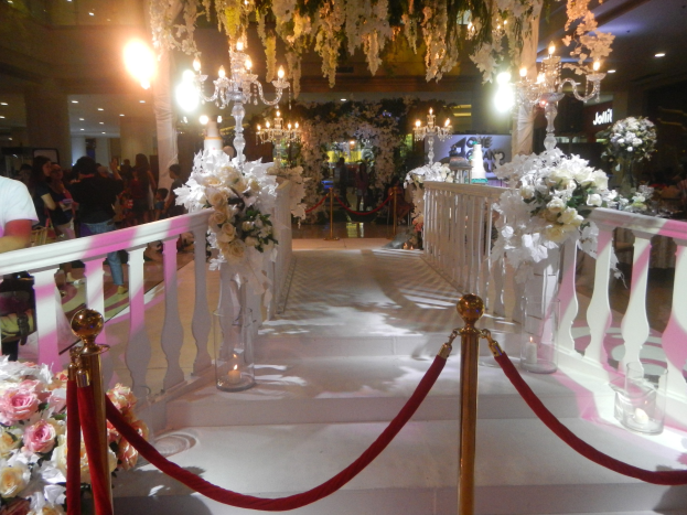 Eine Hochzeitszeremonie in einem Einkaufszentrum mit weißem Teppich, dekorativen Geländern, Blumenarrangements und Deckenbeleuchtung, bei der eine Menge Menschen mit Taschen anwesend sind.