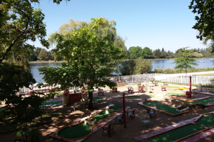 Minigolf-Platz in einem Park mit verstreuten Stühlen und Tischen, umgeben von Bäumen und einem Zaun, mit einem Gewässer und einem Boot im Hintergrund unter dem Himmel.
