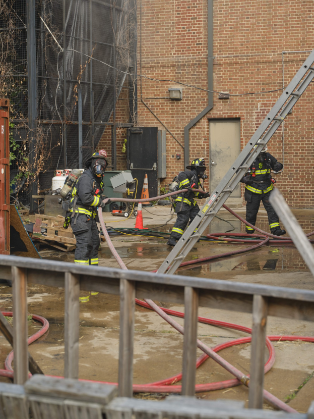 Feuerwehrleute in Helmen arbeiten daran, ein Gebäude Feuer zu löschen, während sie Schläuche halten, mit einem Metallzaun, Rohren, einem Container, einer Verkehrskegel, Gegenständen, Fenstern, Tür, Metallrahmen, Drähte, Baum und Himmel sichtbar.
