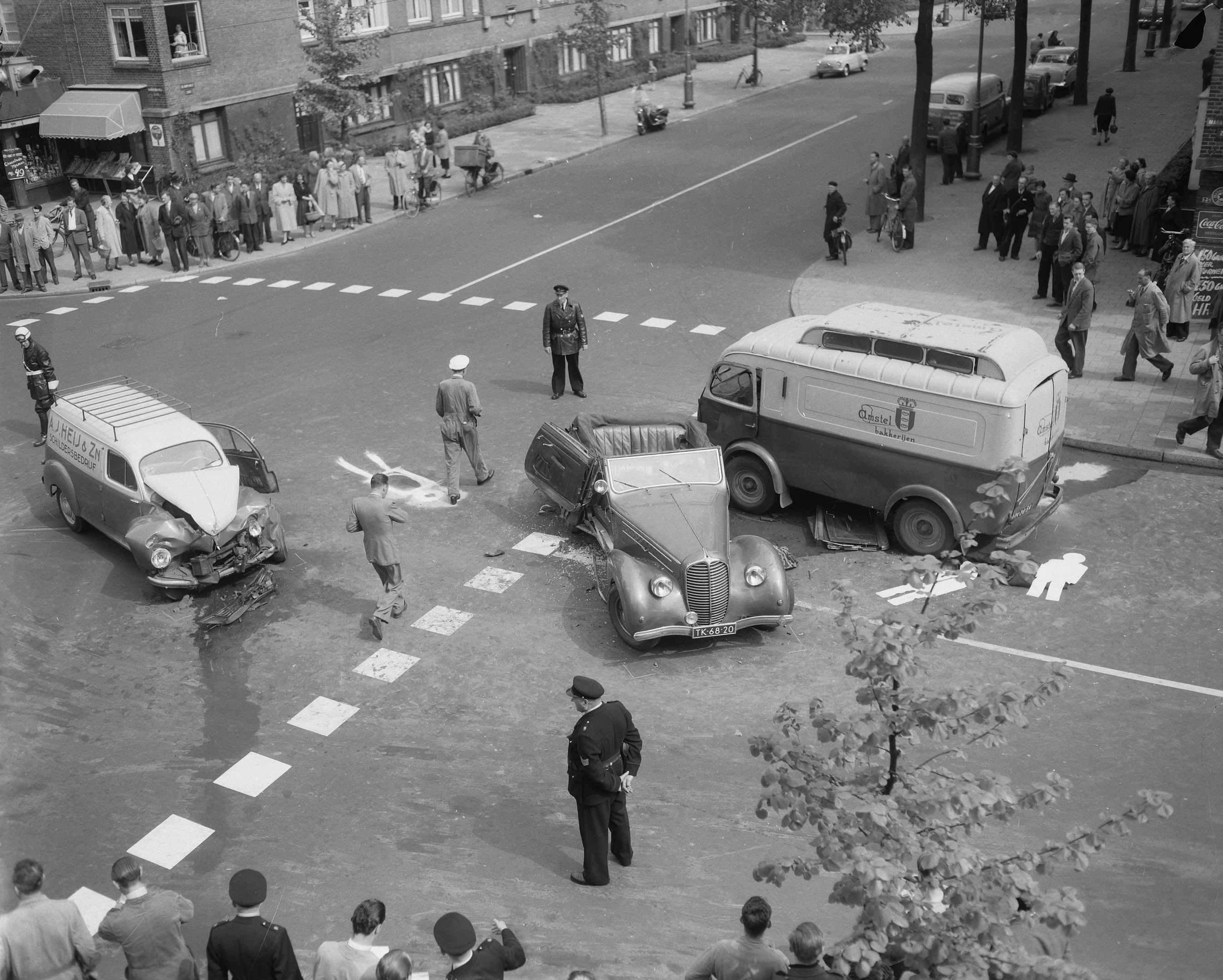 Schwarz-weißes Bild eines Autounfalls an einer Straßenkreuzung mit mehreren Fahrzeugen, darunter ein Lieferwagen, und einer Gruppe von Schaulustigen; Gebäude, Bäume, Pfosten und Schilder sind im Hintergrund zu sehen.