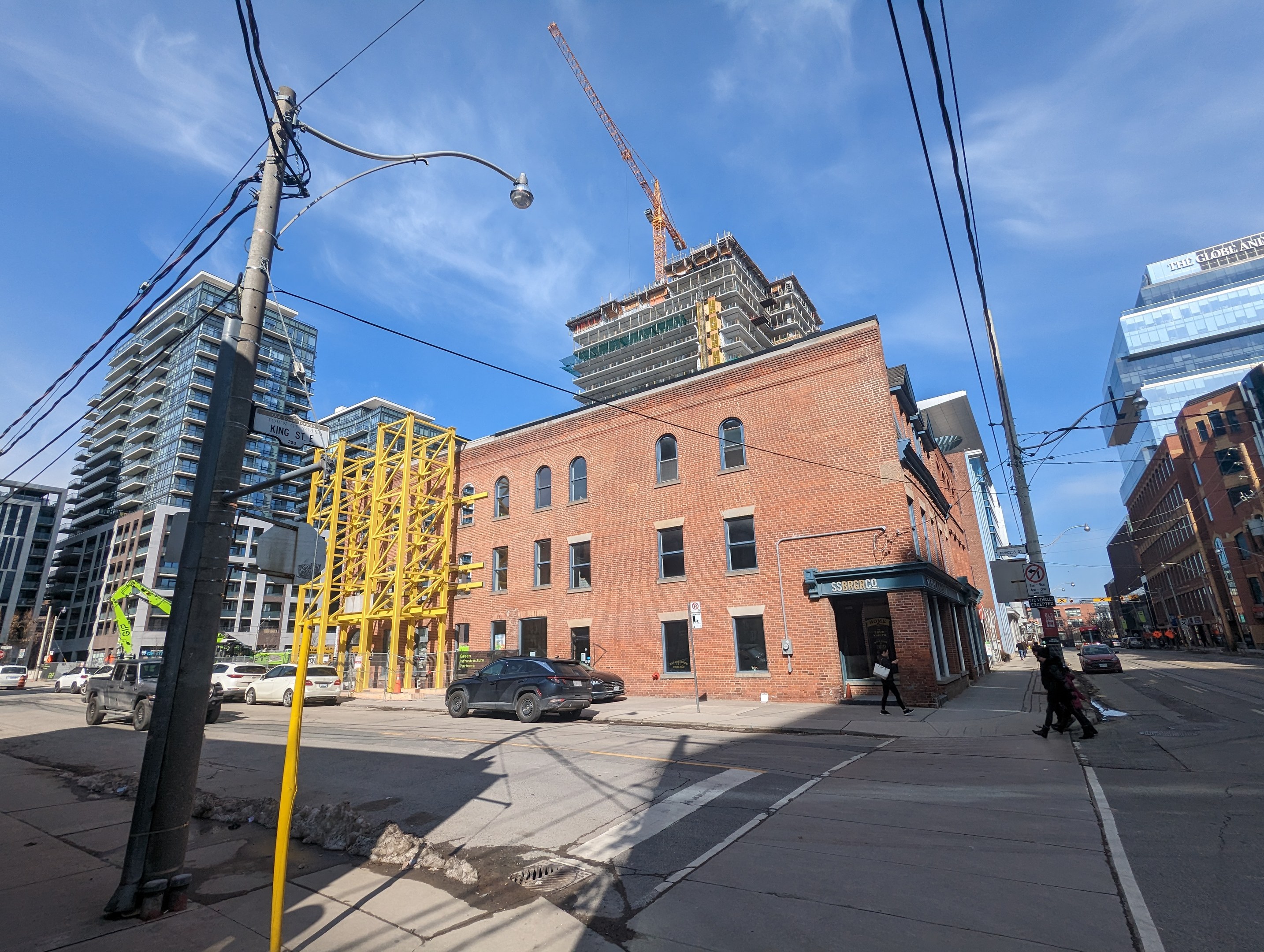 Eine belebte Stadtstraße mit fahrenden Fahrzeugen, Fußgängern auf dem Gehweg, Strommasten, Straßenlaternen und Gebäuden, die einen Baukran am Himmel zeigen.