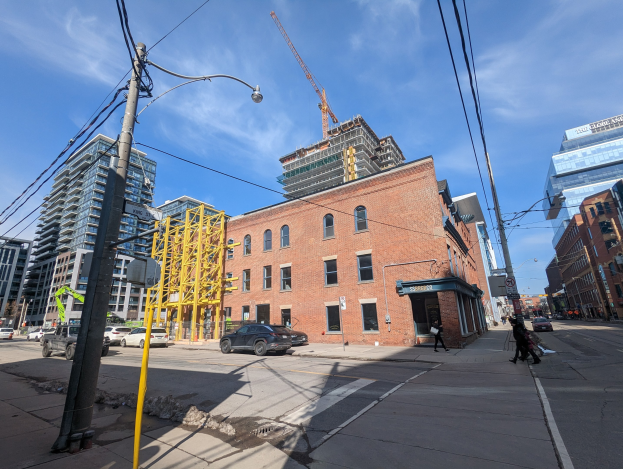 Eine belebte Stadtstraße mit fahrenden Fahrzeugen, Fußgängern auf dem Gehweg, Strommasten, Straßenlaternen und Gebäuden, die einen Baukran am Himmel zeigen.