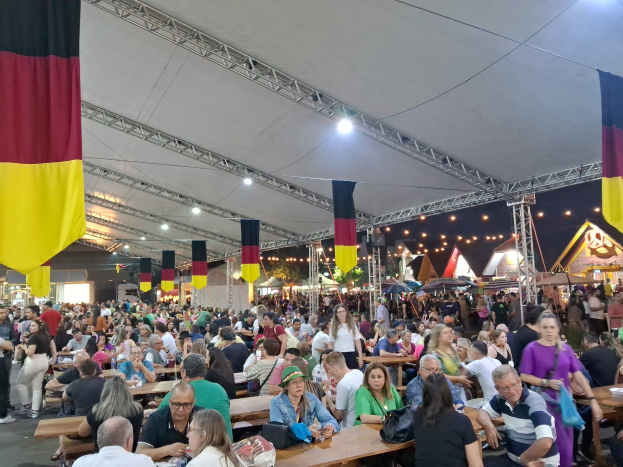 Eine große Gruppe von Menschen, die an Tischen unter einem Zelt auf dem Oktoberfest in München sitzen, mit einigen, die stehen und Taschen auf den Tischen, und Zelten, Lichtern und Fahnen im Hintergrund.