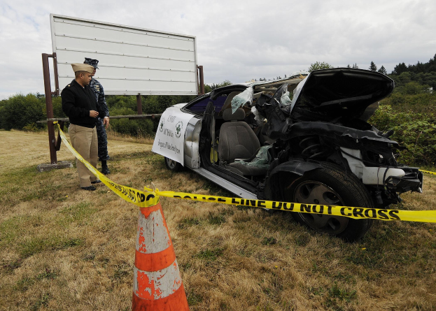 Ein Polizist in Uniform steht neben einem schwer beschädigten Auto am Straßenrand, mit eingedellter Motorhaube und zersplitterten Fenstern, neben einem Verkehrskegel und einem Band an einem Pfahl, mit Bäumen und einem bewölkten Himmel im Hintergrund.